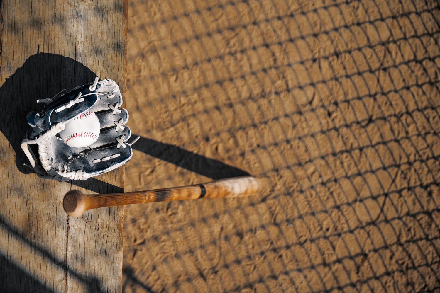 Baseball bat and baseball in a baseball mit sitting on a bat on the right side of image places aesthetically. Angle from the top. Red dirt in the immediate background blurred. 